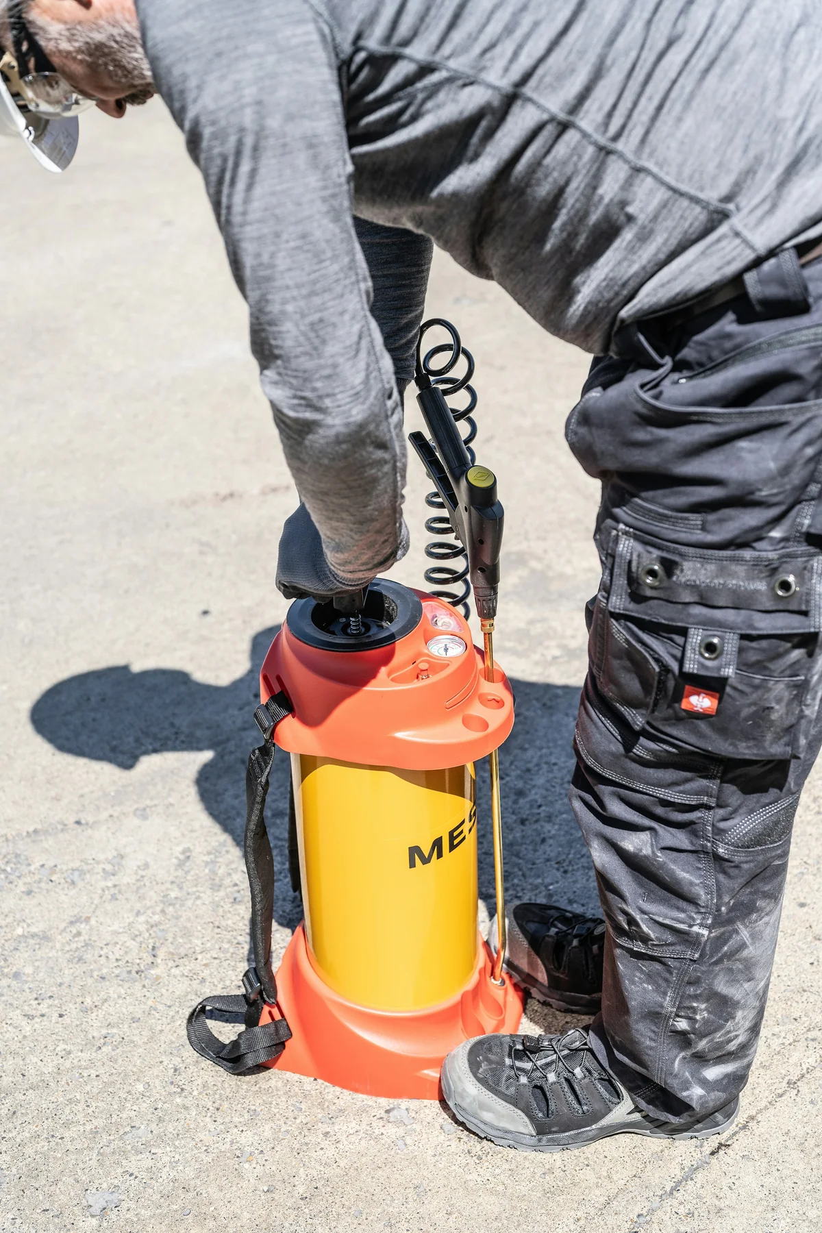 A gloved civilian worker preparing a yellow and orange pressure sprayer